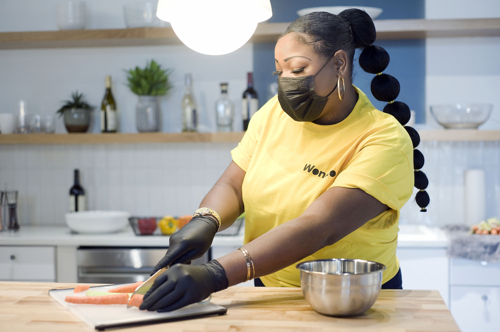 A woman wearing a mask and gloves cuts carrots in a kitchen during her gig work.