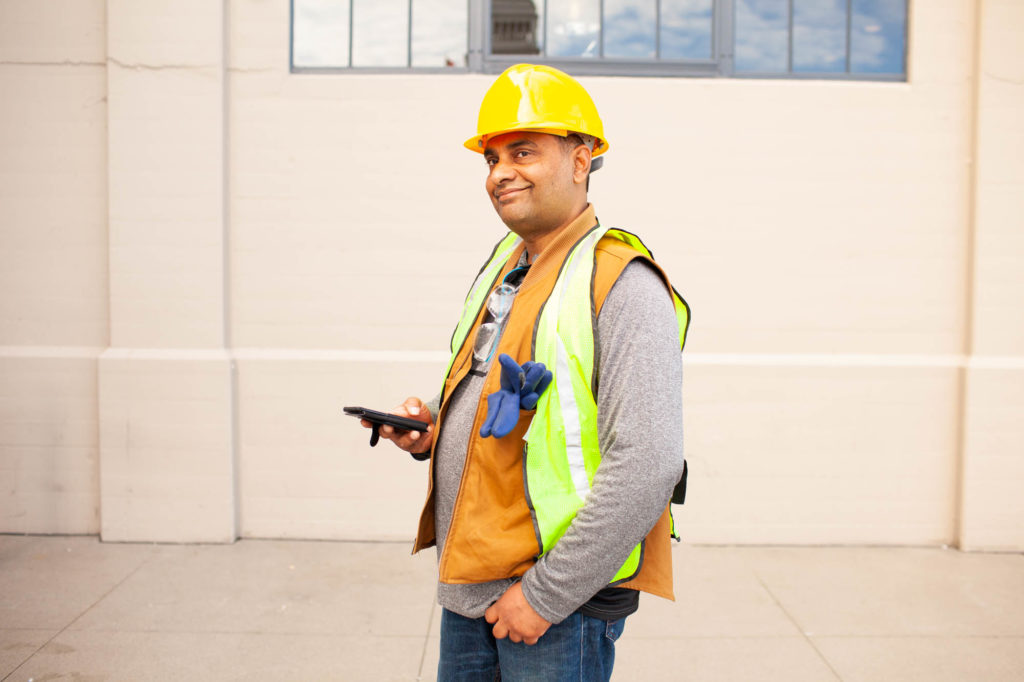 A man in a hard hat stands outside his warehouse job ready to look for other available jobs on his phone.