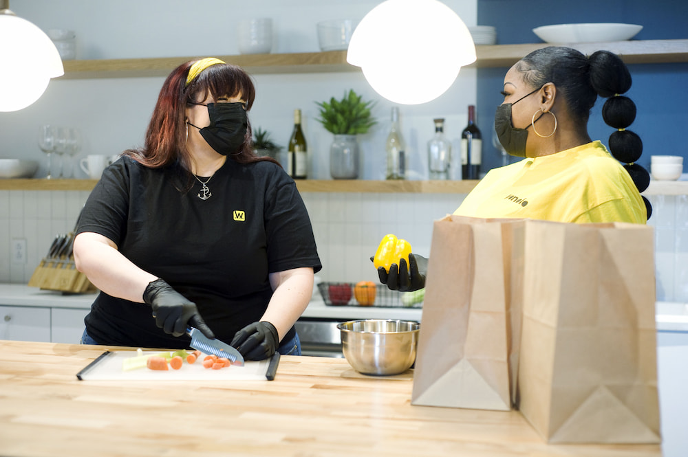 Two women in a kitchen are wearing masks and gloves while cutting fruits and vegetables during their part-time side hustle.