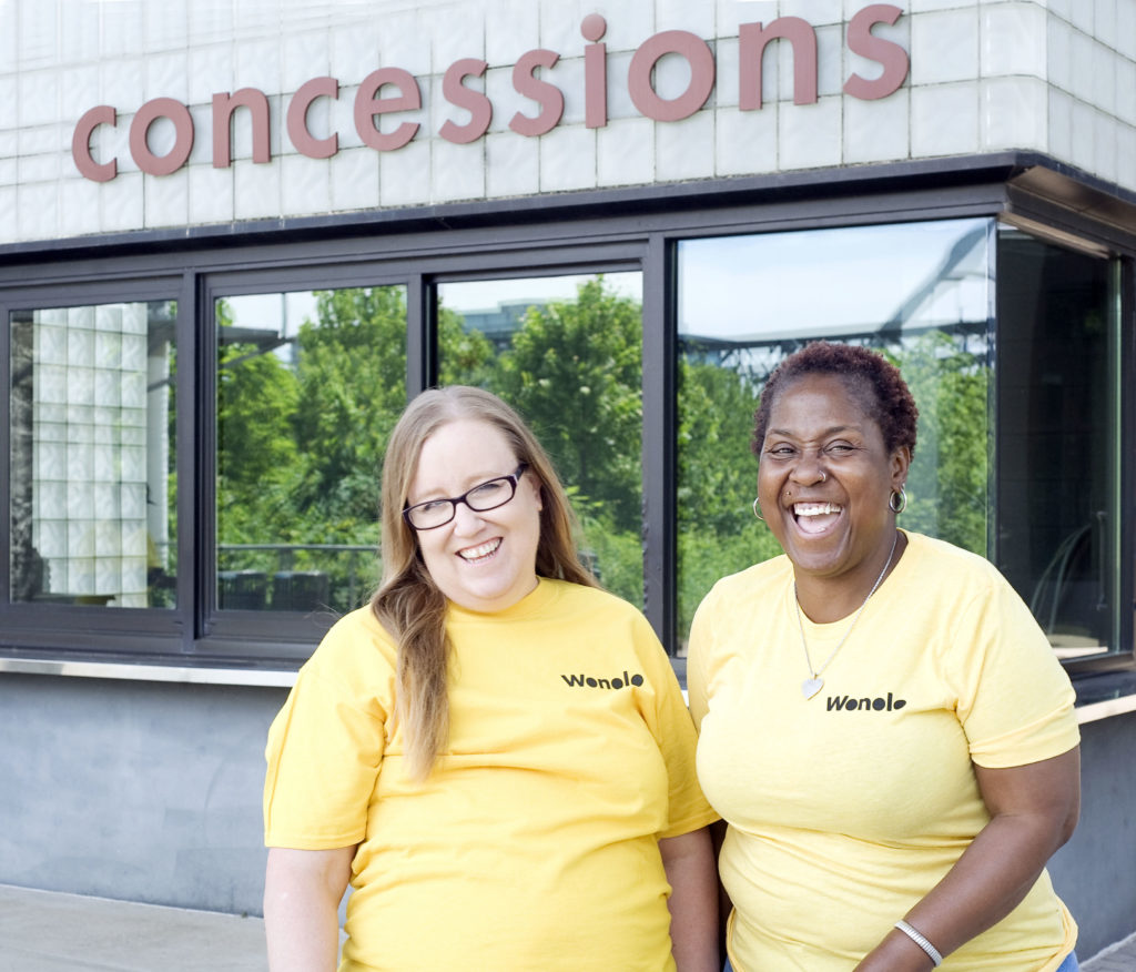 Two workers in yellow Wonolo shirts stand smiling in front of a concession stand.