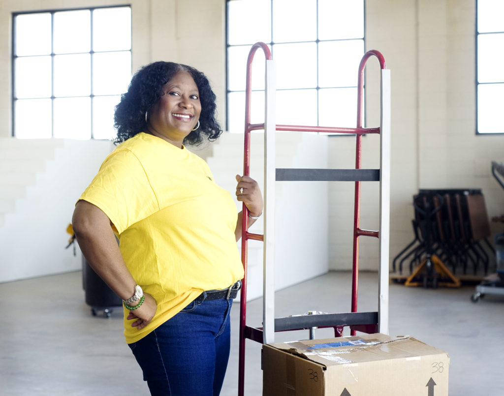 A temporary worker in a yellow Wonolo shirt is standing next to a moving dolly with a box on it.